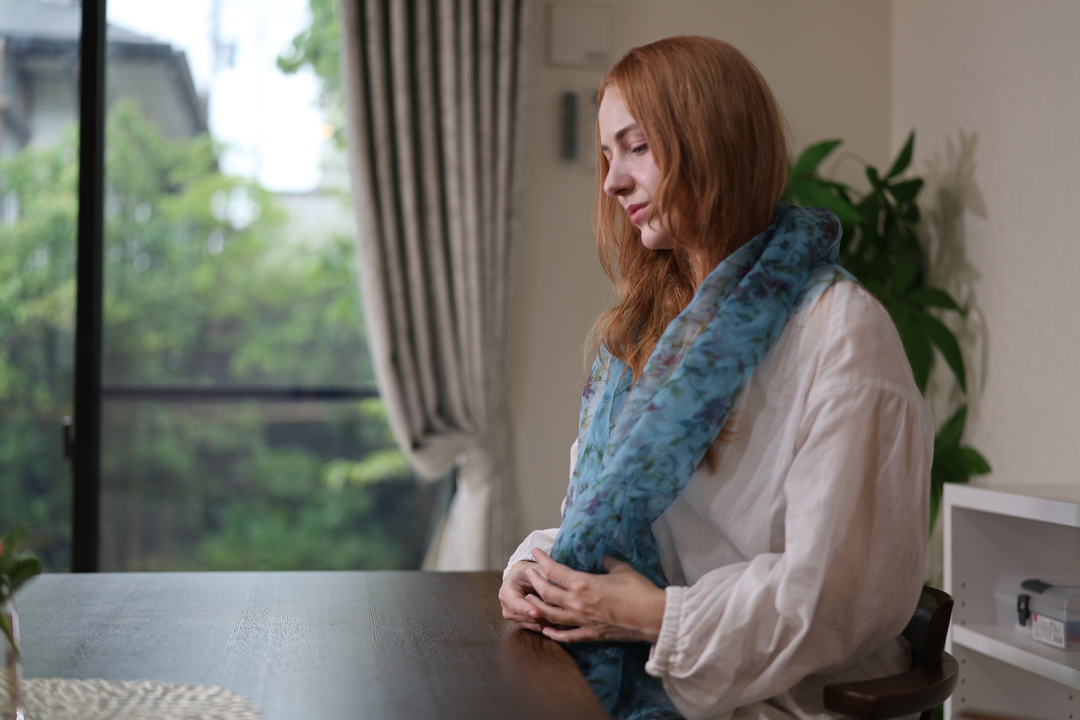 A seated model wearing the blue silk stole with both hands resting together at a dark table.