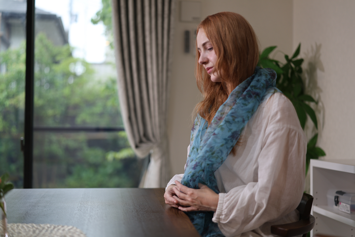 A seated model wearing the blue silk stole with both hands resting together at a dark table.