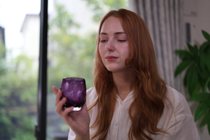 A seated model pausing with the Gold-Purple HYOUSYOU Edo Kiriko glass in one hand beside a bright window.