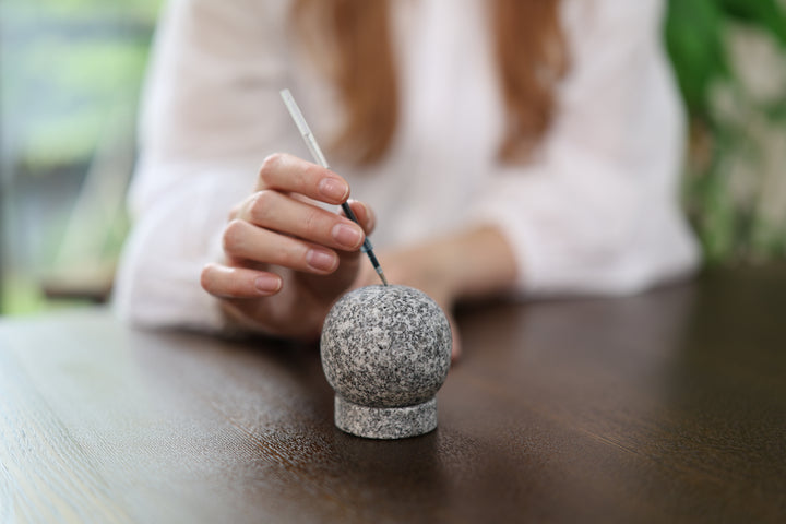 A model bringing an unlit incense stick to the Makabe Stone Incense Stone on a broad wooden table.