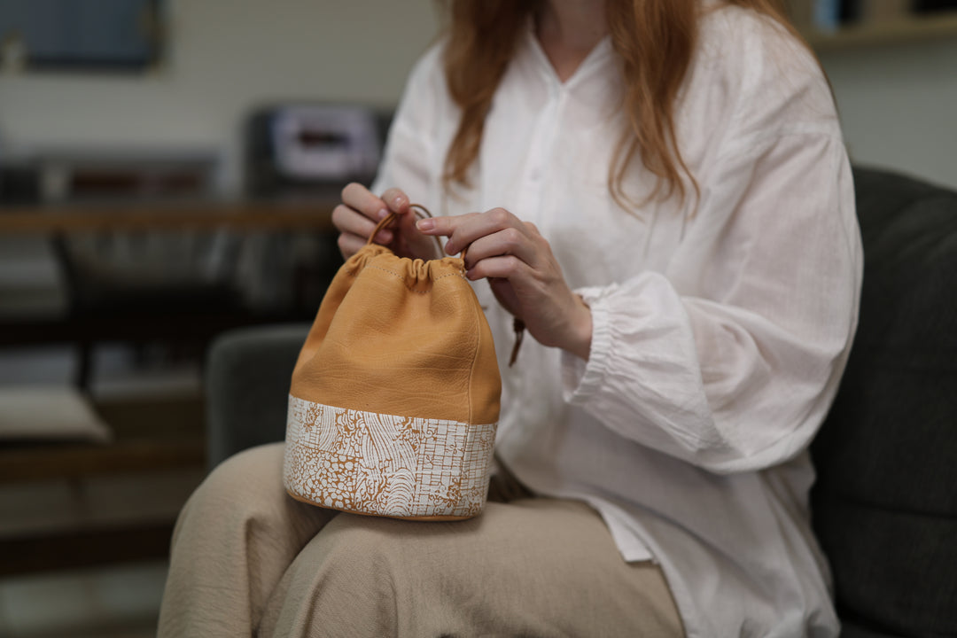 A seated model on a sofa adjusting the cords of the natural leather Edo Komon pouch on her lap.
