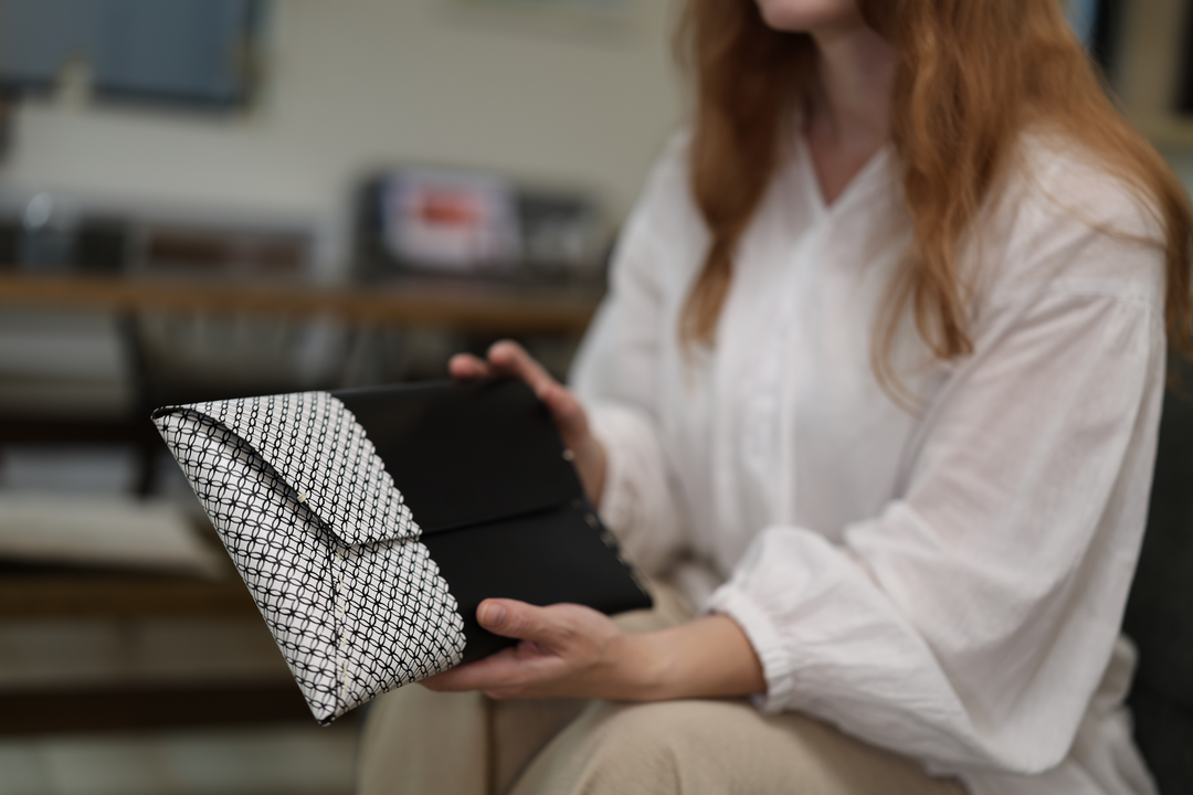 A seated model opening the black leather and white Tokyo Some-Komon clutch beside a café table.