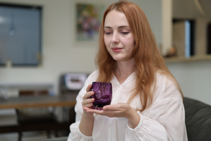 A seated model looking at the Gold-Purple HYOUSYOU Edo Kiriko glass while holding it with both hands on a sofa.
