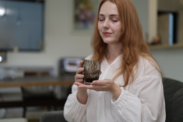 A seated model holding the HYOUSYOU Blue Ink Edo Kiriko glass in a relaxed living-room setting.