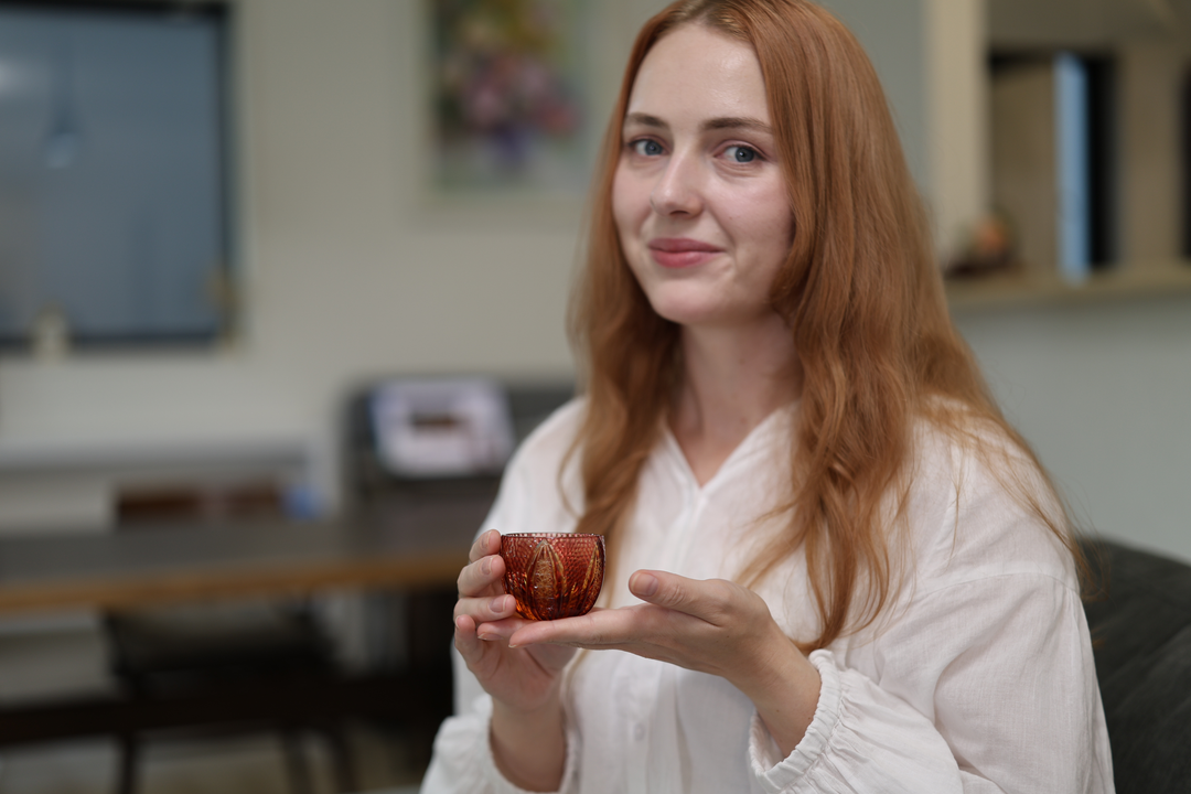 Adult model seated on a sofa presenting the GENEN Gold-Red and Amber Edo Kiriko glass between both hands.