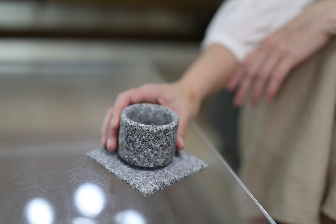 A seated model steadying a stone cup on the Makabe Stone Art Plate on a reflective glass table beside a sofa.