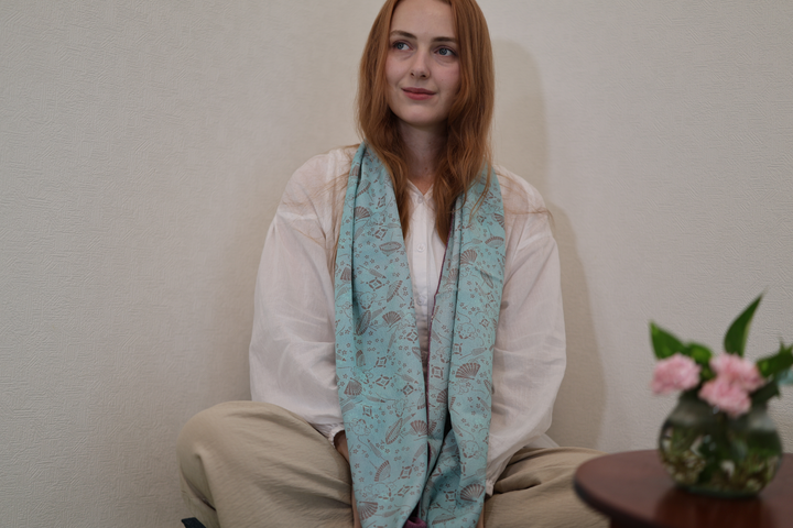 A seated model wearing the long pale blue face of the double-sided silk stole in a quiet Japanese room.