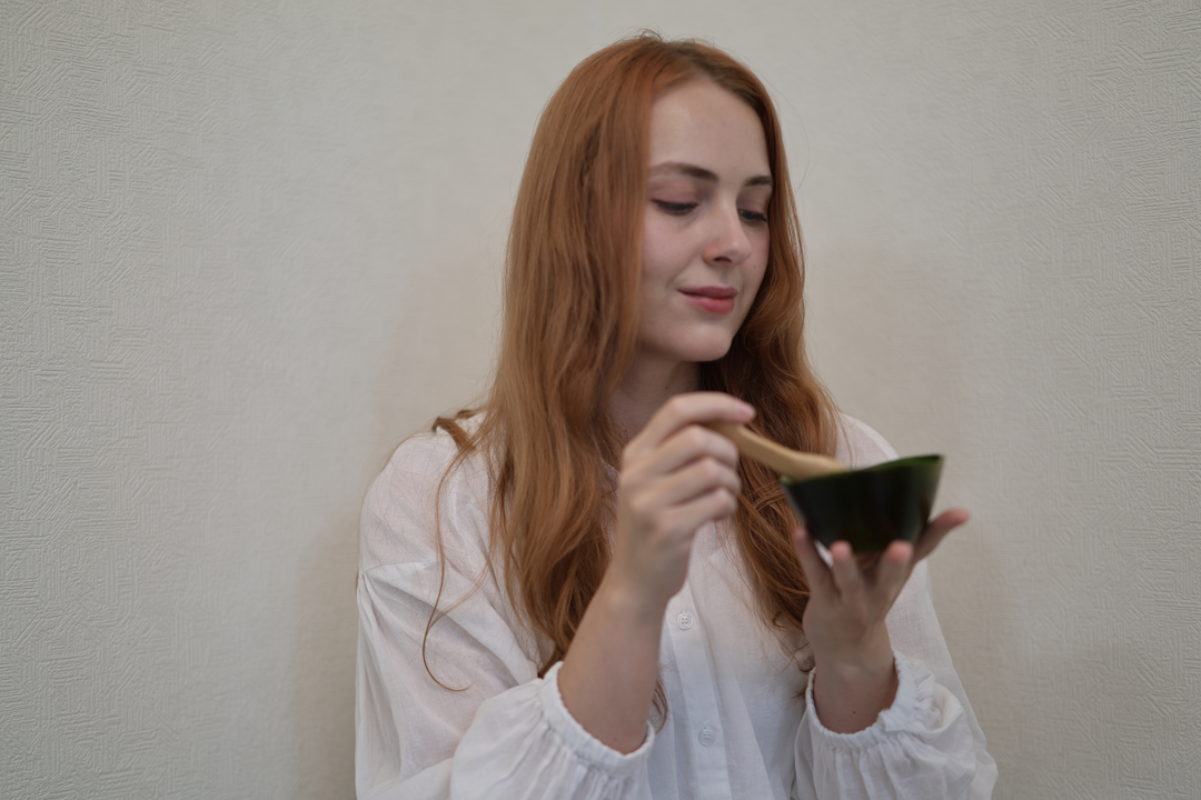 Adult model seated at a red table, holding RIN Bowl S Olive in one hand and a wooden spoon in the other.