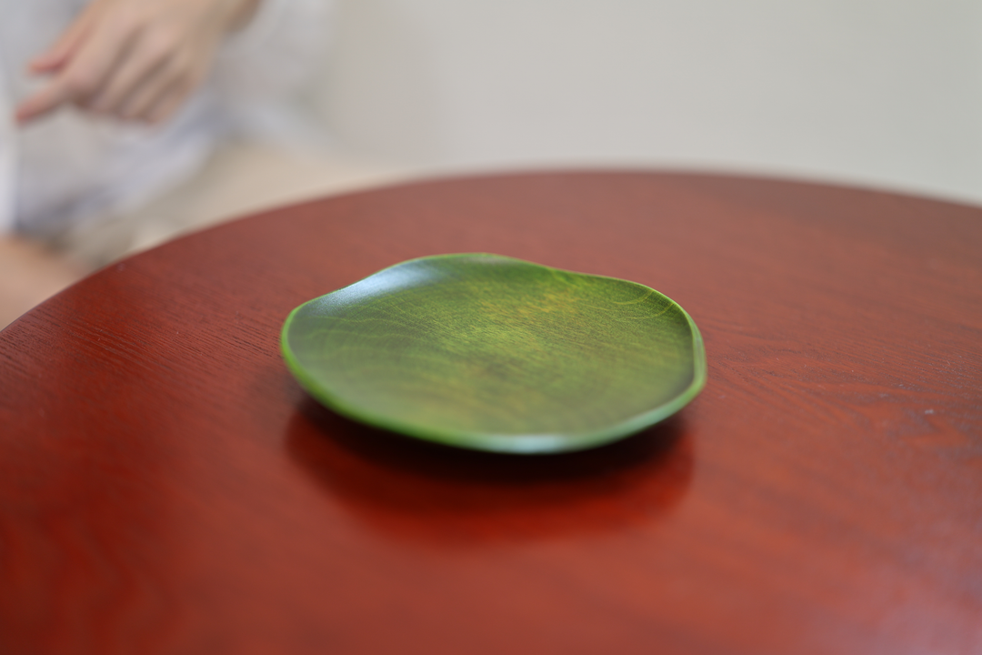 RIN 5.0 Olive plate resting on a red-brown table with a person's hand softly out of focus behind it.