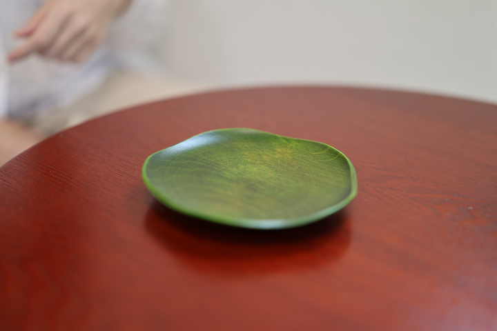RIN 5.0 Olive plate resting on a red-brown table with a person's hand softly out of focus behind it.