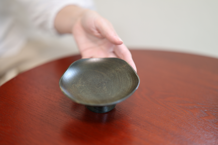A hand reaching toward an empty Floating Leaf Sesame on a red-lacquered table.