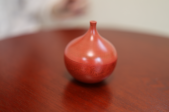The closed berry-red Bulb lacquerware vessel on a wooden table with a seated model softly visible behind it.