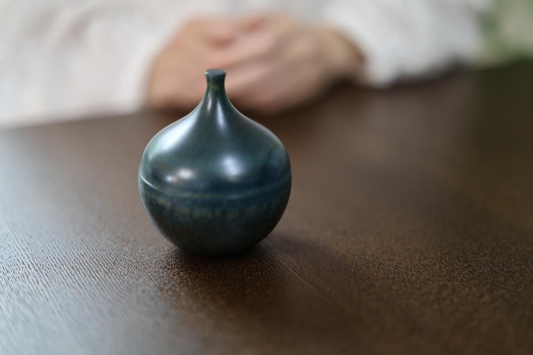 Bulb: Hyacinth standing closed on a dark table before a seated model with both hands at rest.