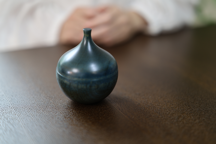 Bulb: Hyacinth standing closed on a dark table before a seated model with both hands at rest.