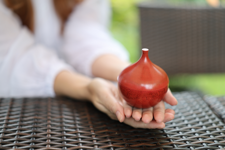 A model presenting the closed berry-red Bulb lacquerware vessel on an open palm beside an outdoor table.
