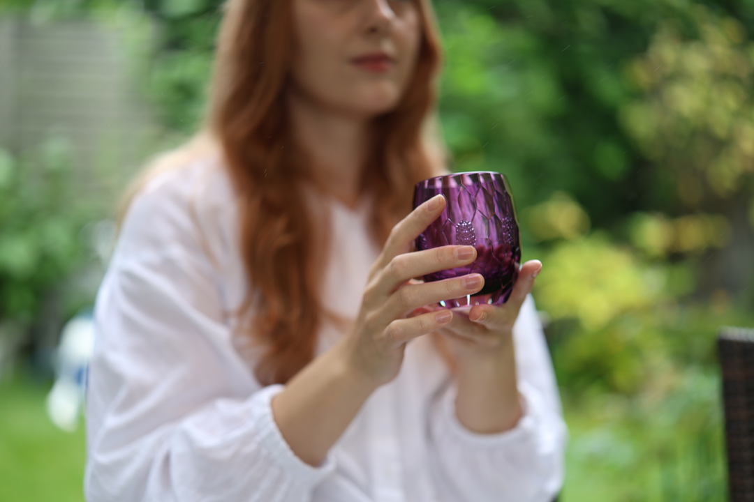 A model holding the Gold-Purple HYOUSYOU Edo Kiriko glass securely in both hands against green garden foliage.