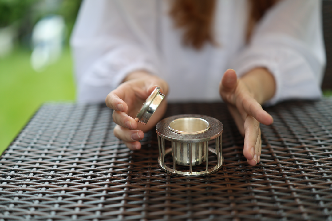 A model lifting the vented lid from the pure silver incense burner at a woven outdoor table.