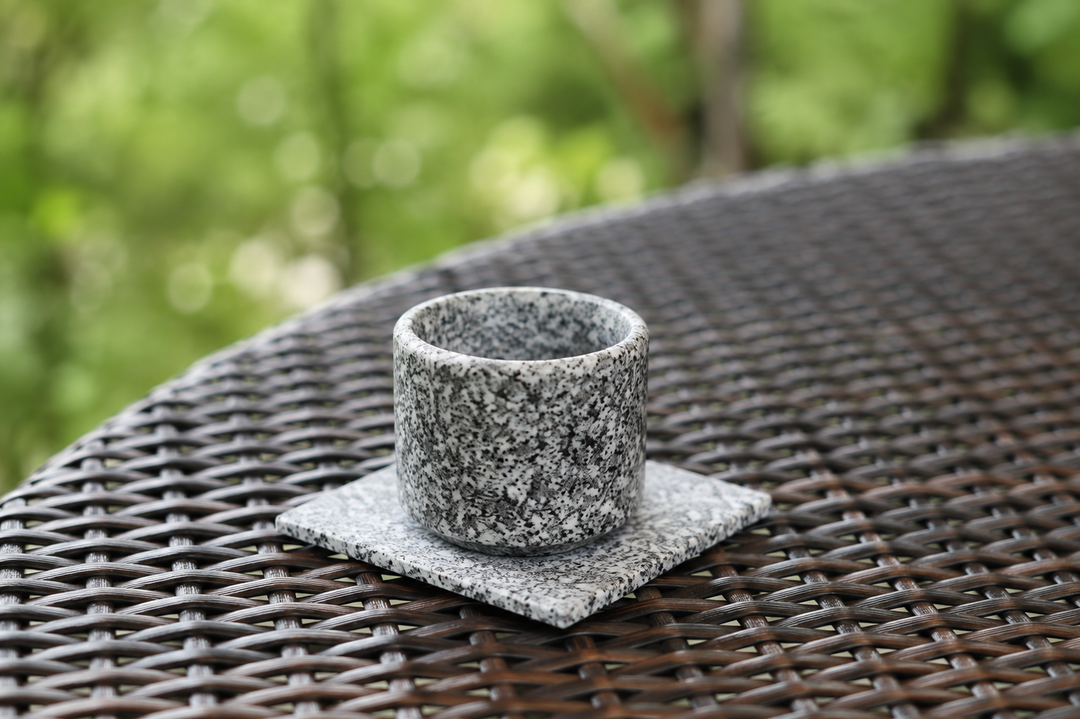 A cup on the square Makabe Stone Art Plate on an outdoor woven table in gentle natural light.
