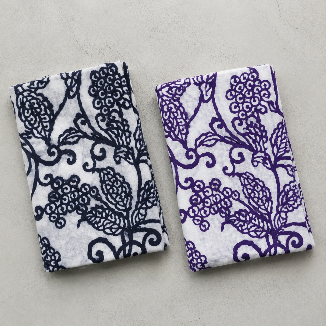 Two folded tenugui colorways placed side by side indoors to compare the mood of the shared arabesque pattern.