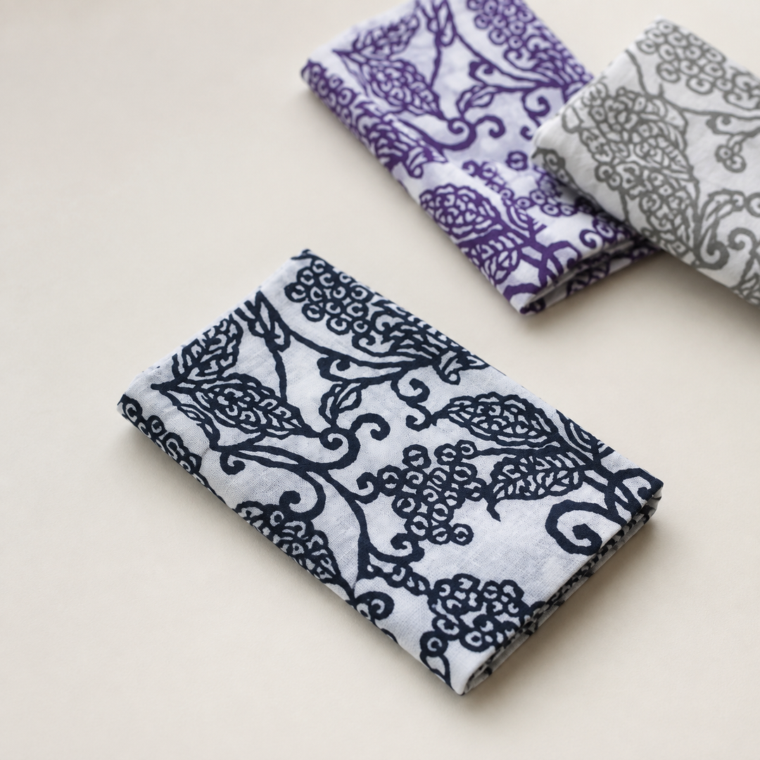 A dark-blue arabesque-pattern tenugui given quiet space on a plain indoor surface, with two companion colorways behind.