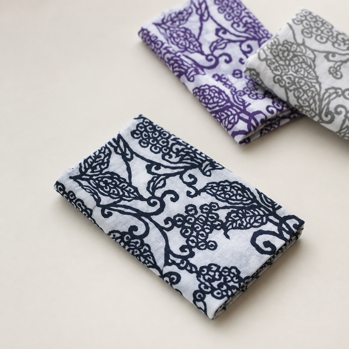 A dark-blue arabesque-pattern tenugui given quiet space on a plain indoor surface, with two companion colorways behind.