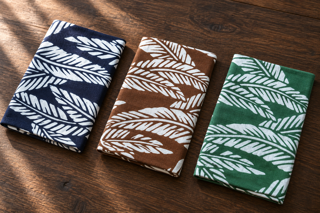 Three folded Basho tenugui in navy, brown, and green arranged on a wooden surface with the white leaf motifs visible.