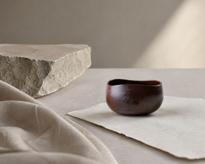 Bud: Lilac lacquerware vessel beside matte paper, pale stone, and woven cloth.