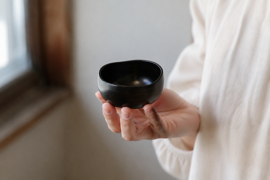 The small sesame-black Bud lacquerware vessel held close to a window, showing its gently irregular rim.