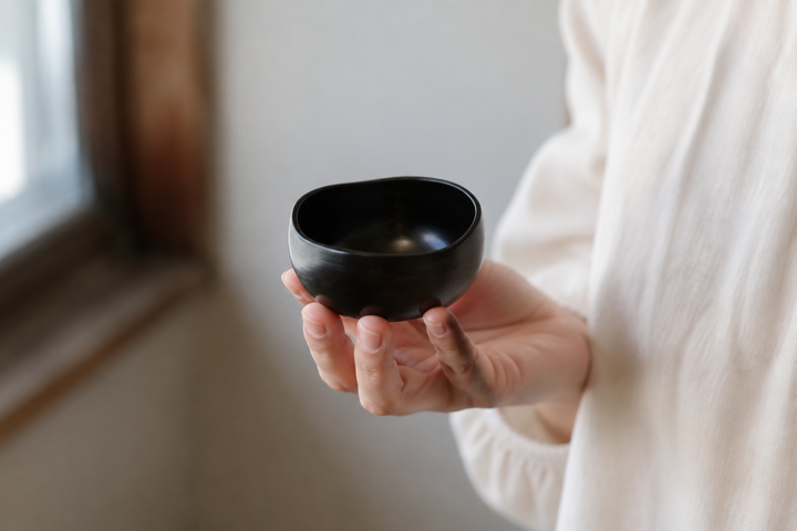 The small sesame-black Bud lacquerware vessel held close to a window, showing its gently irregular rim.
