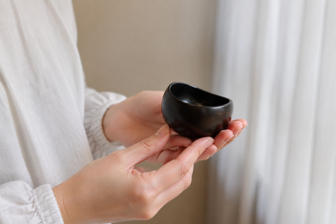 Two hands gently rotating the empty sesame-black lacquerware vessel beside a softly lit window.