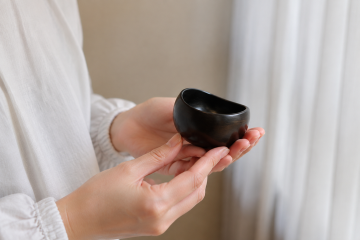 Two hands gently rotating the empty sesame-black lacquerware vessel beside a softly lit window.