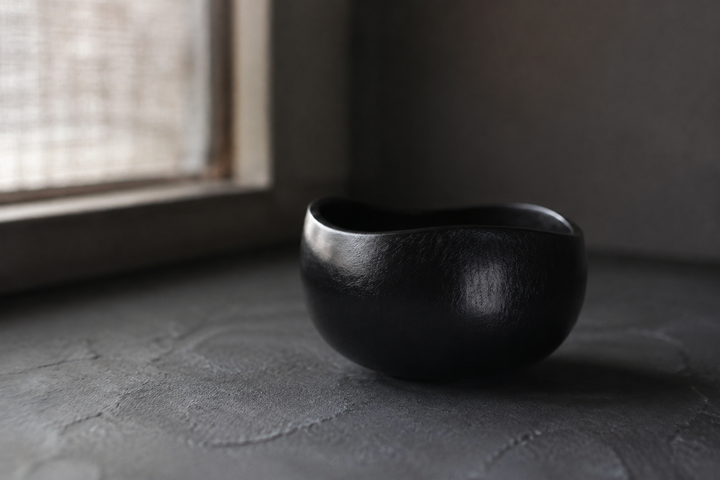 Soft window light tracing the curved dark lacquer surface of the small vessel.