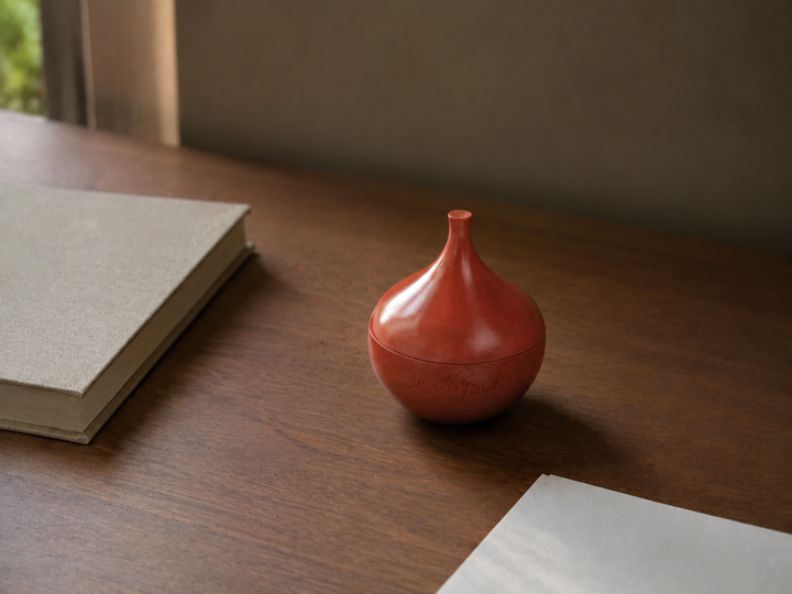 The closed berry-red Bulb lacquerware object beside a book and paper on a spare reading desk.
