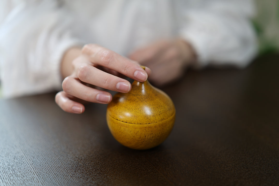 A hand lightly touching the tip of the closed Bulb: Mimosa on a dark table before lifting the lid.