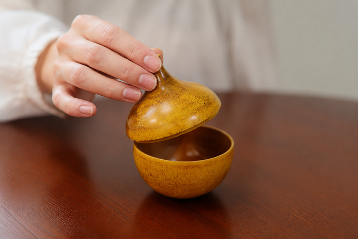 An adult hand gently returning the dry Bulb: Mimosa lid to its lower section on a clean wooden table.