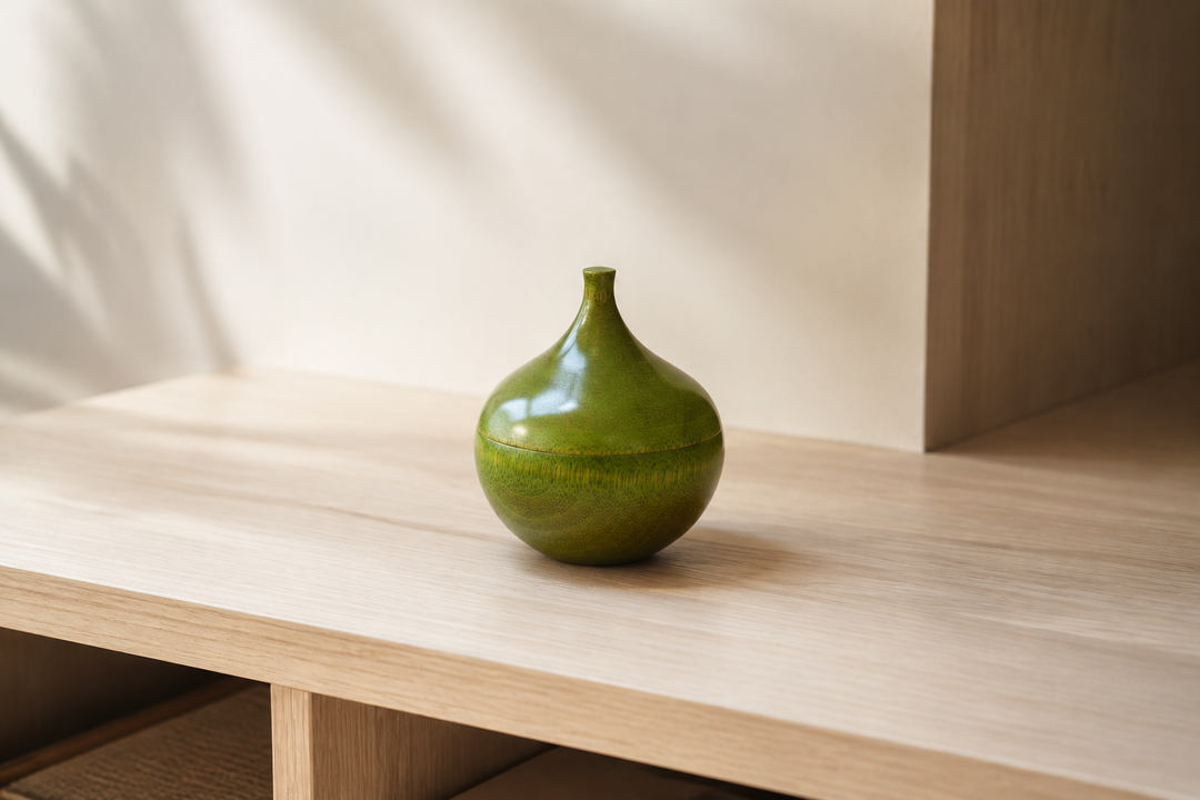 The closed olive-green Bulb lacquerware object standing alone on a quiet pale-wood shelf in soft daylight.