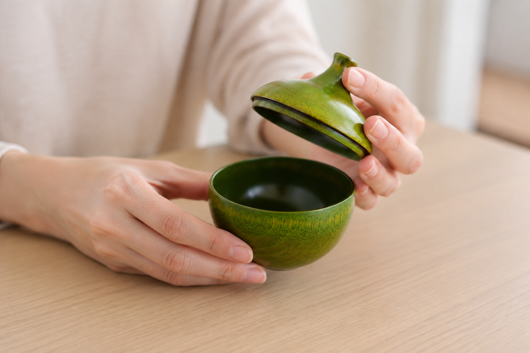 Dry adult hands gently lifting the pointed olive lacquer lid above its matching small bowl on a calm table.