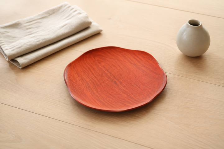 Berry-red RIN 7.0 lacquerware plate beside undyed linen, pale wood, and a small matte ceramic object.