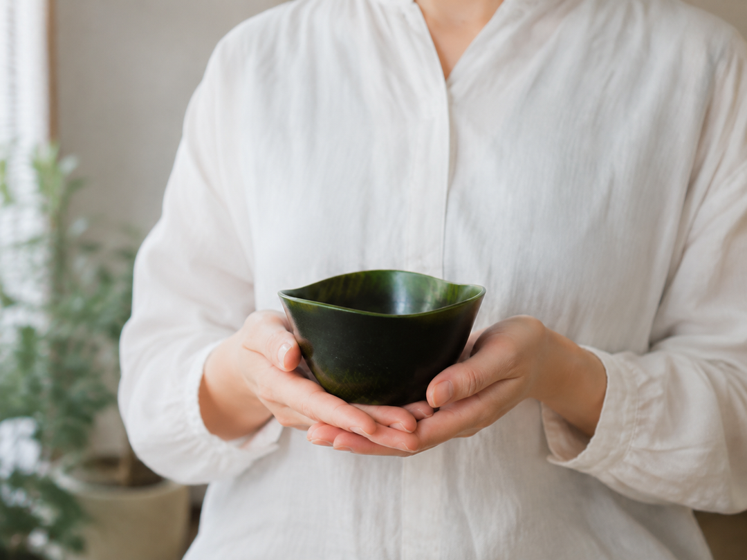 Adult hands gently supporting the empty RIN Bowl M Olive from below, showing its 120-millimeter scale and rising rim.