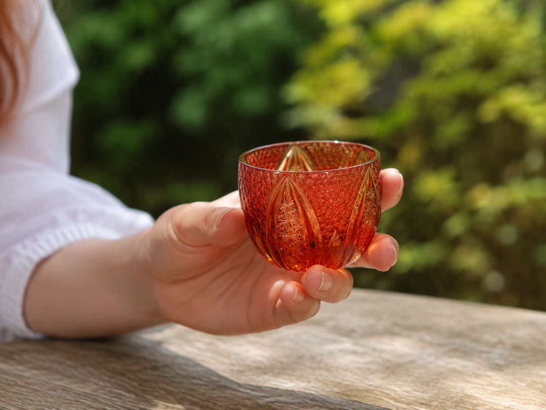 GENEN Gold-Red and Amber glass held above an outdoor table against softly blurred greenery.