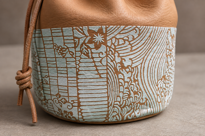 Close view of the pale blue-and-white Edo Komon patterned band, leather grain, seam, and drawstring knot on the natural pouch.