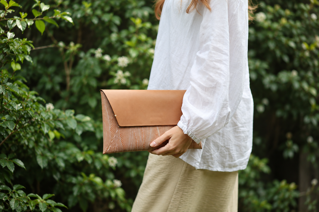 The natural leather Edo Komon clutch carried beside deep green garden foliage in soft outdoor light.