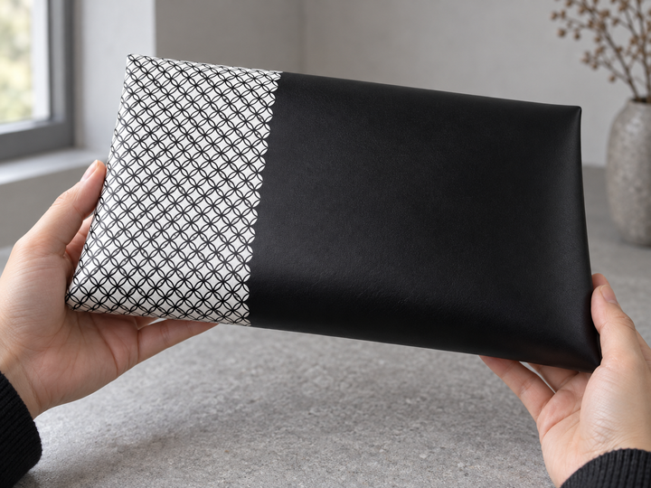 A close three-quarter view of the clutch being turned by hand, showing the fine black repeat wrapping around the white panel into smooth black leather.
