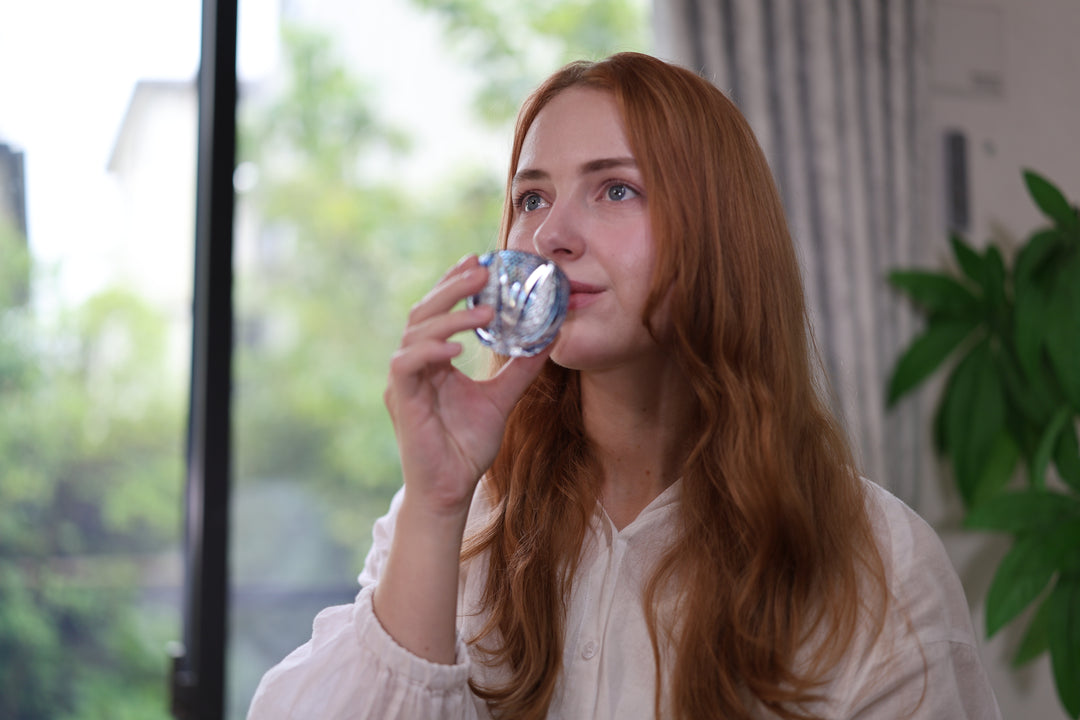 Adult drinking from the GENEN Indigo cut-glass vessel beside a window.