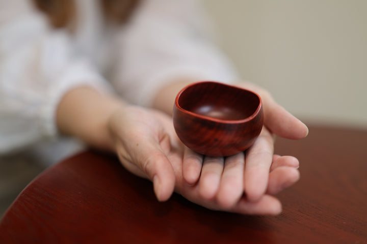 A small berry-red lacquerware vessel resting in a person's open hands above a wooden table.