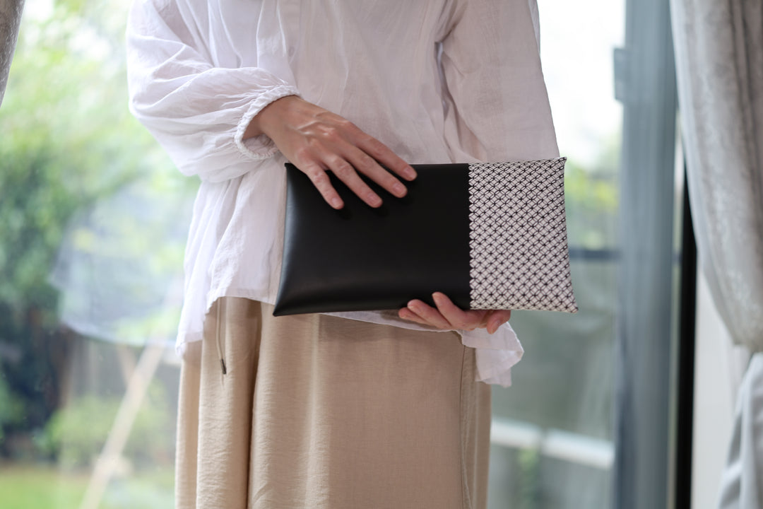 A person holding the black leather and patterned white clutch across both hands beside a bright window.