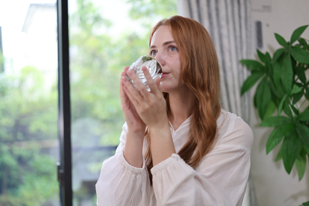 A woman drinking from the Korohana Edo Kiriko vessel beside a bright window.