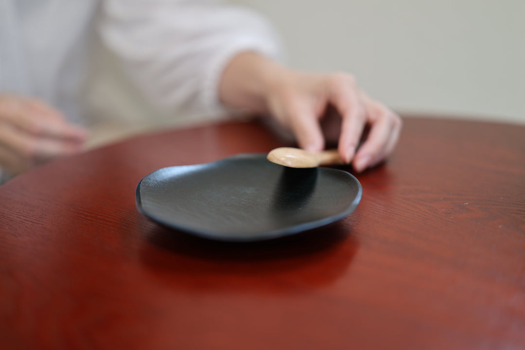 A wooden spoon approaching the rim of the dark lacquered RIN 5.0 Hyacinth plate on a red wooden table.