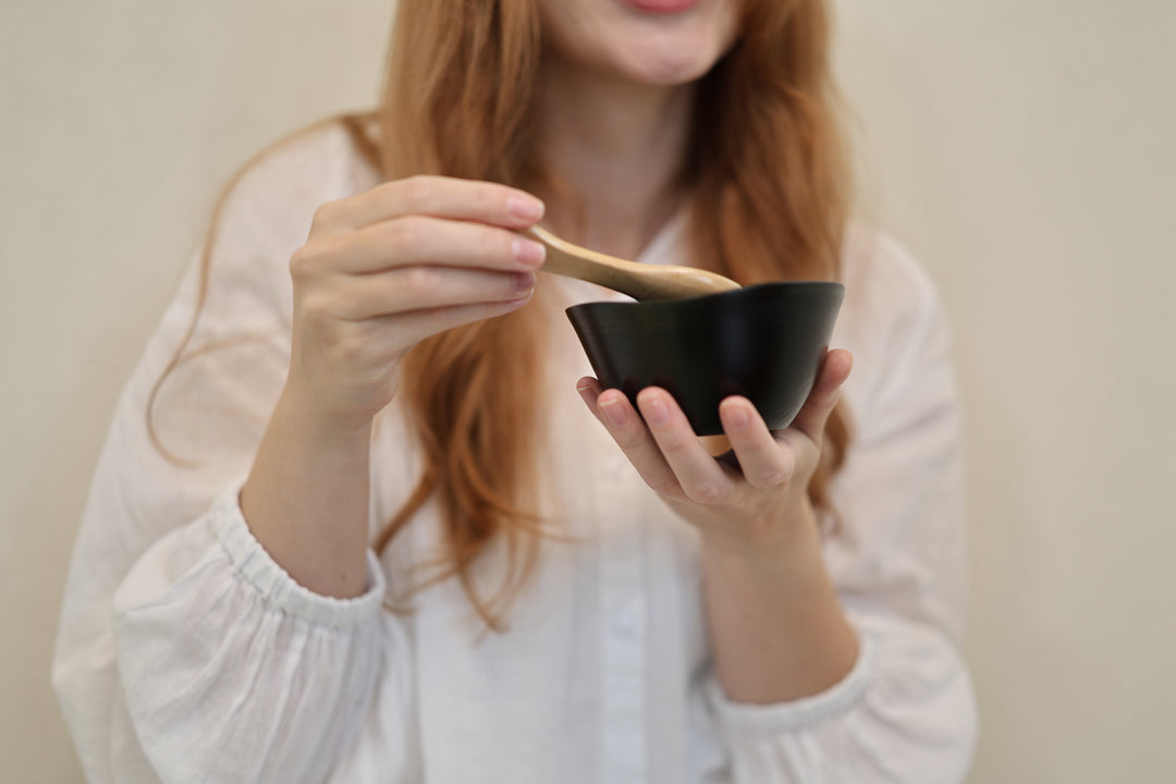 Adult model supporting the empty RIN Bowl M Sesame while holding a wooden spoon above its irregular rim.