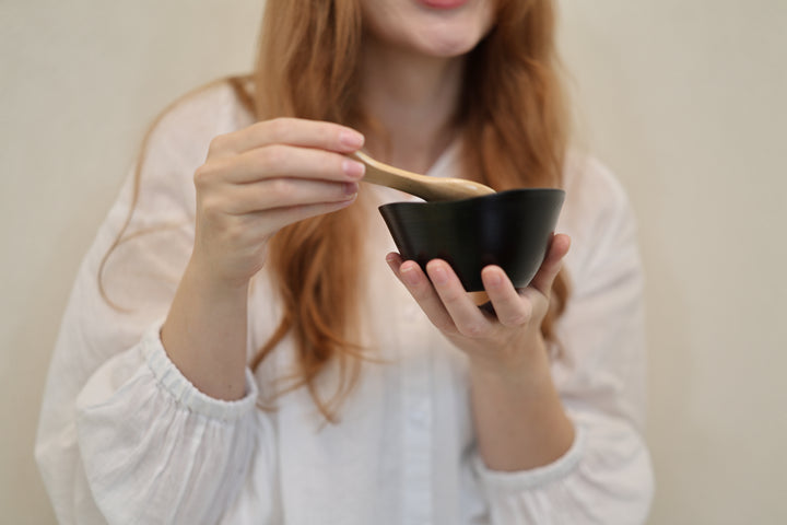 Adult model supporting the empty RIN Bowl M Sesame while holding a wooden spoon above its irregular rim.
